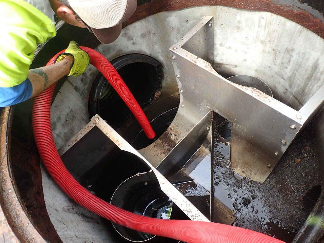 A worker in a high-visibility vest uses a red suction hose to clean sediment from a metal stormwater treatment unit.
