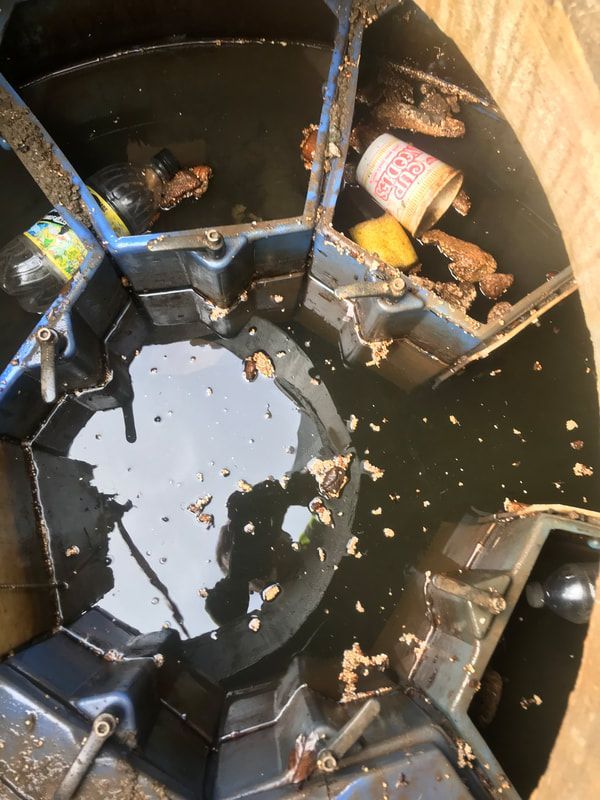 A high-angle view into a storm drain with dark water, discarded plastic trash, a paper cup, and debris.