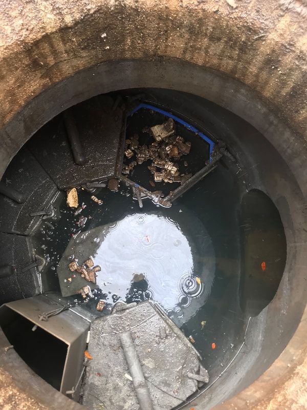 View looking down into a concrete manhole containing stagnant water, debris, and metal equipment components.