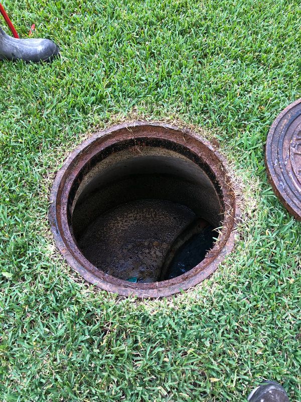An open, circular sewer or utility access manhole set into a patch of green grass, with its metal lid resting nearby.