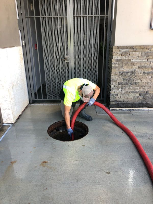 A worker in a high-visibility yellow shirt and blue gloves uses a large red hose to clean a circular drain in the ground.