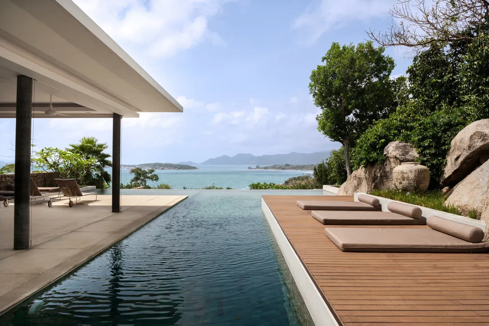 Infinity pool overlooking ocean, modern villa with wooden deck and lounge chairs, blue sky.