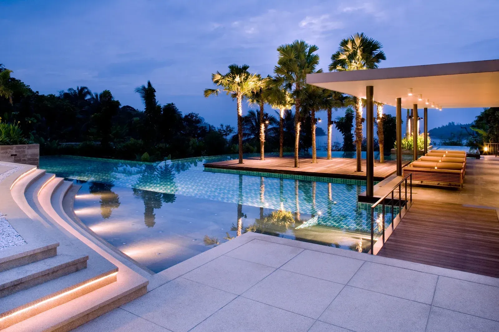 Luxury resort pool at dusk, with seating under a canopy and palm trees. Calm water reflects the sky.