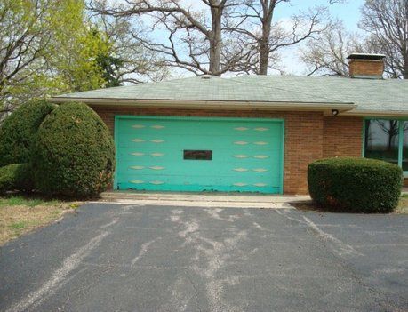Garage with a teal door in front of a brick house, flanked by green bushes; driveway in foreground.