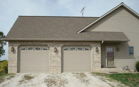 Two-car garage with tan doors and light brick, driveway, attached to a tan house with a brown roof.
