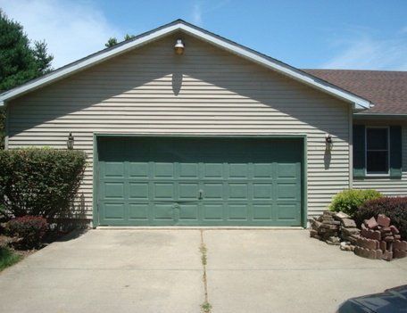 Green garage door with light tan siding, concrete driveway, and landscaping.