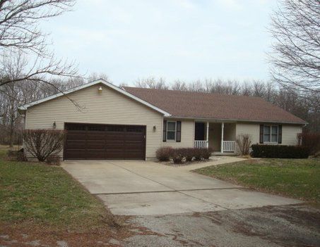Tan ranch-style house with a brown garage door and driveway, set against a background of trees.