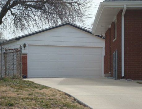 White garage with closed door, brick house beside it, driveway, and chain-link fence.