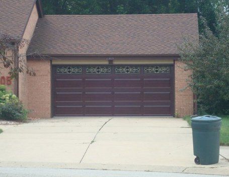 Brown garage door with decorative top panels, brown roof, brick facade, and green trash bin on a concrete driveway.