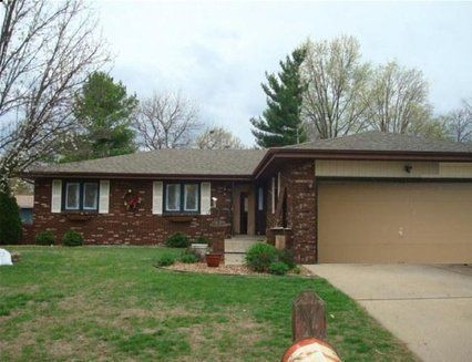 Brick ranch-style house with a tan garage door and green lawn under a cloudy sky.