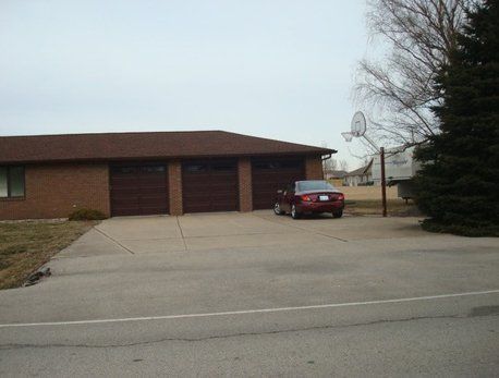 Red car parked in front of a brick building with three garage doors and a basketball hoop.
