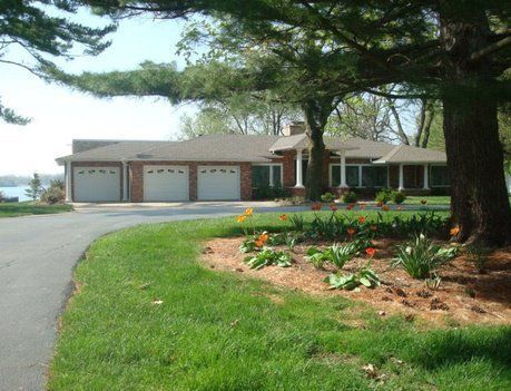 House with a three-car garage, brick exterior, and green lawn; driveway curves towards the home; large tree on the right.