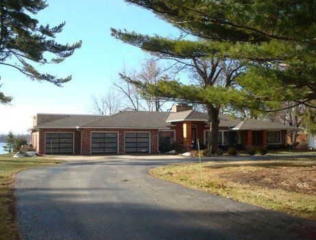 House with a three-car garage, brick facade, and circular driveway, set in a grassy yard with trees under a blue sky.