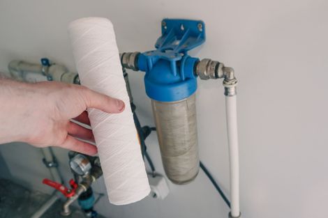 Hand holding a new white water filter next to a water filter system on a wall.