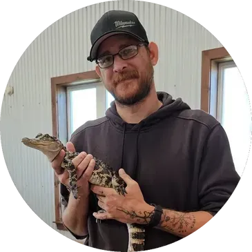 Man holding a small alligator; wearing a cap, glasses, and a hoodie; indoor setting.