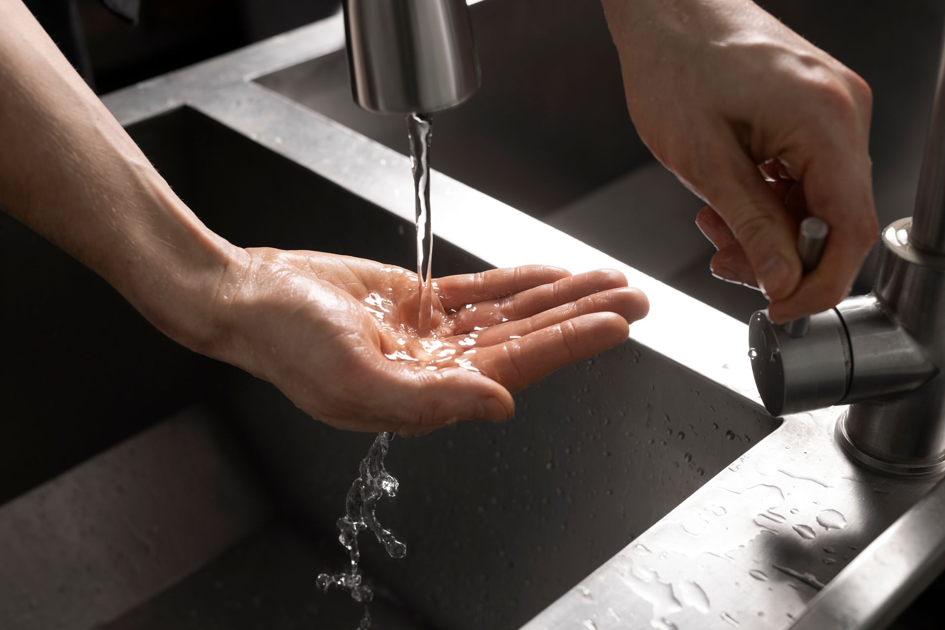 Person's hands under running water from a kitchen faucet, next to a stainless steel sink.