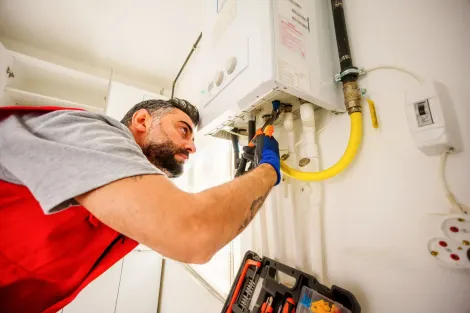 Plumber working on a gas boiler; using tools; wearing gloves. White wall, yellow gas pipe.