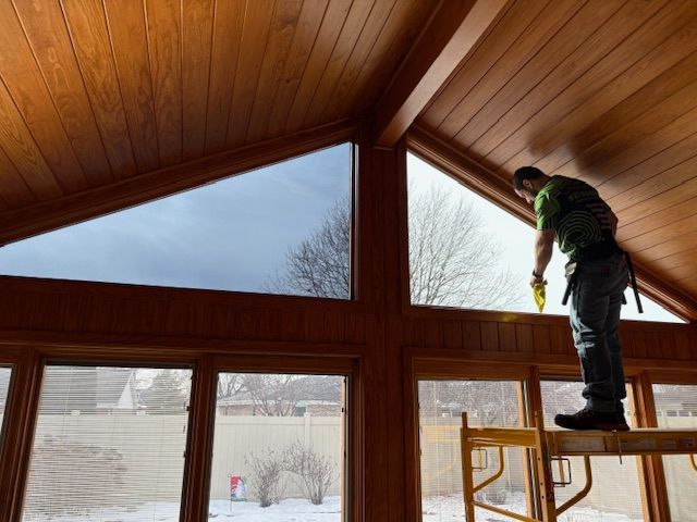 A person window washing in a sunroom with wood paneling and a snowy outdoor view.