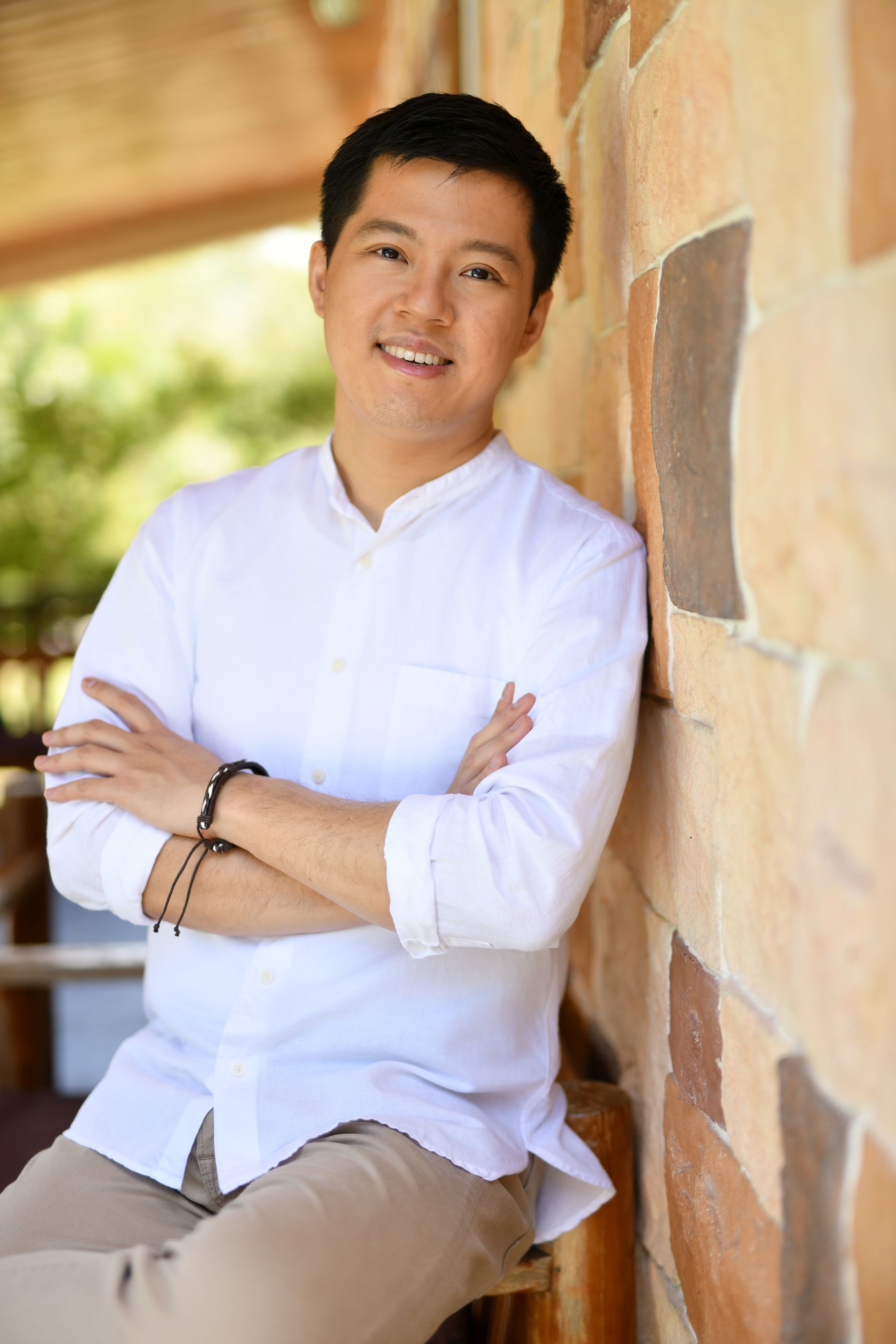 Man in white shirt leans against a textured stone wall, arms crossed, smiling.