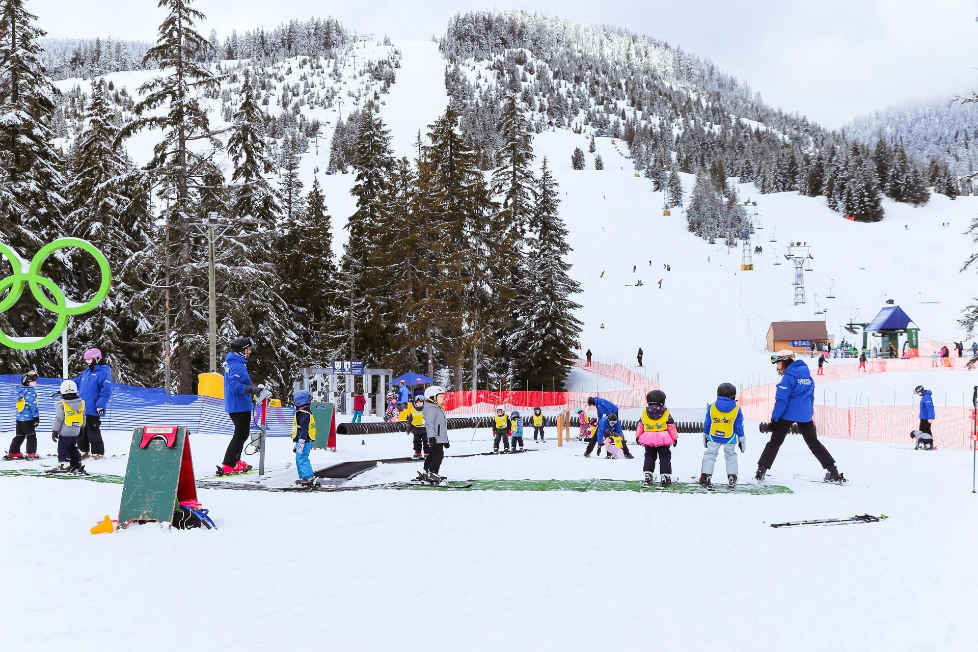 Children learning to ski with instructors on a snowy slope, Olympic rings in the background.