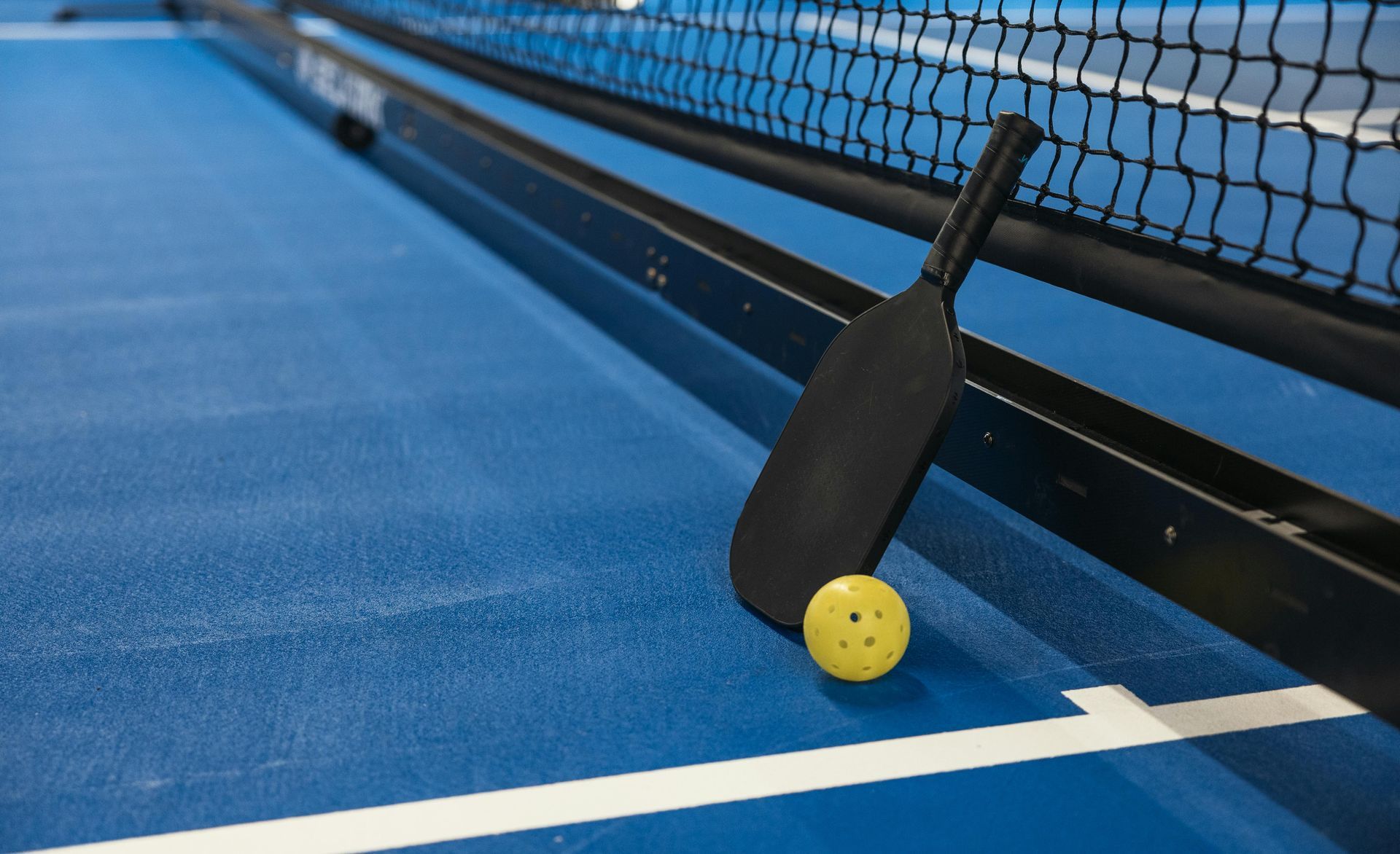 Black pickleball paddle and yellow ball on a blue court next to a net.