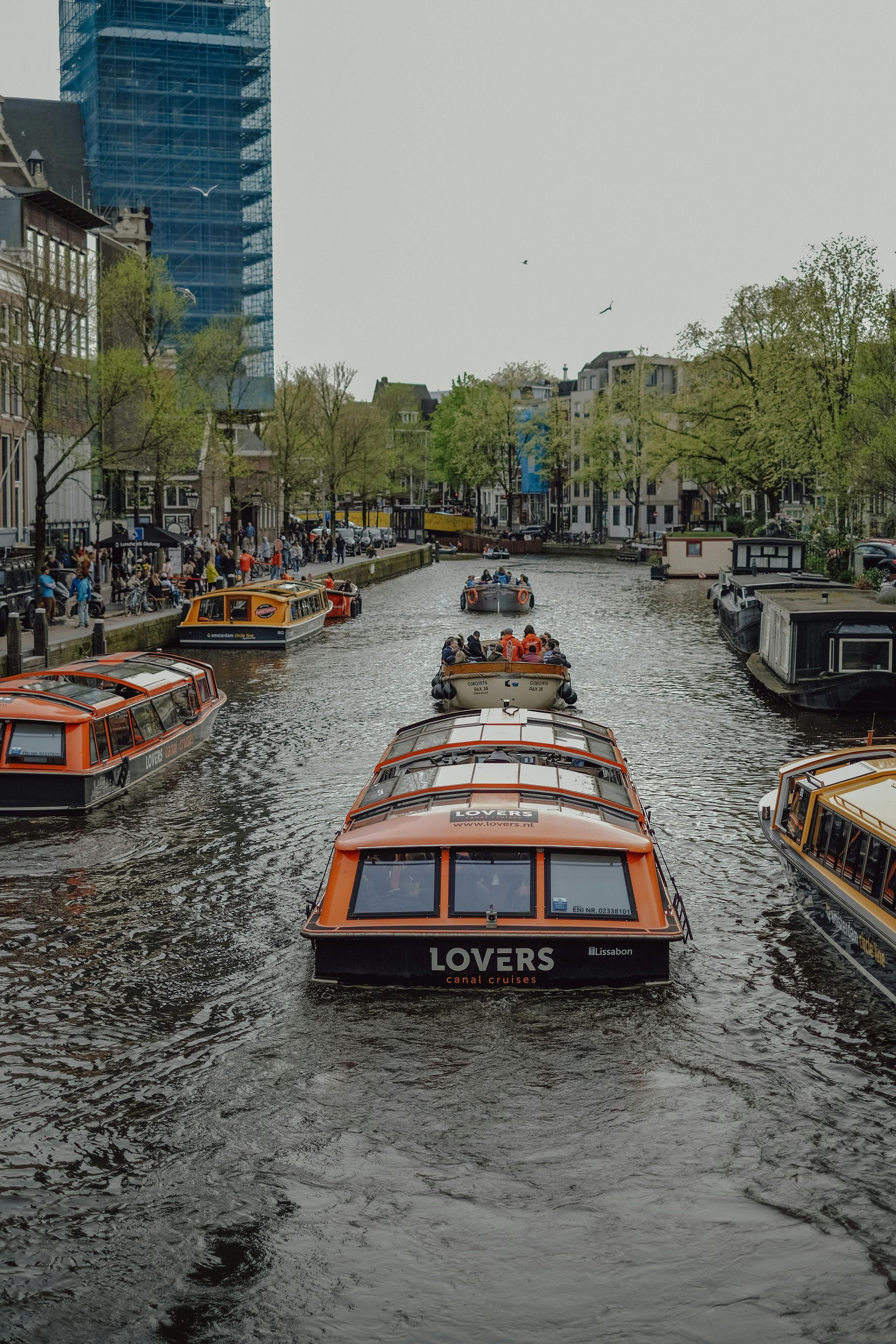 Orange tour boats on canal in Amsterdam, with people, buildings, and cloudy sky.