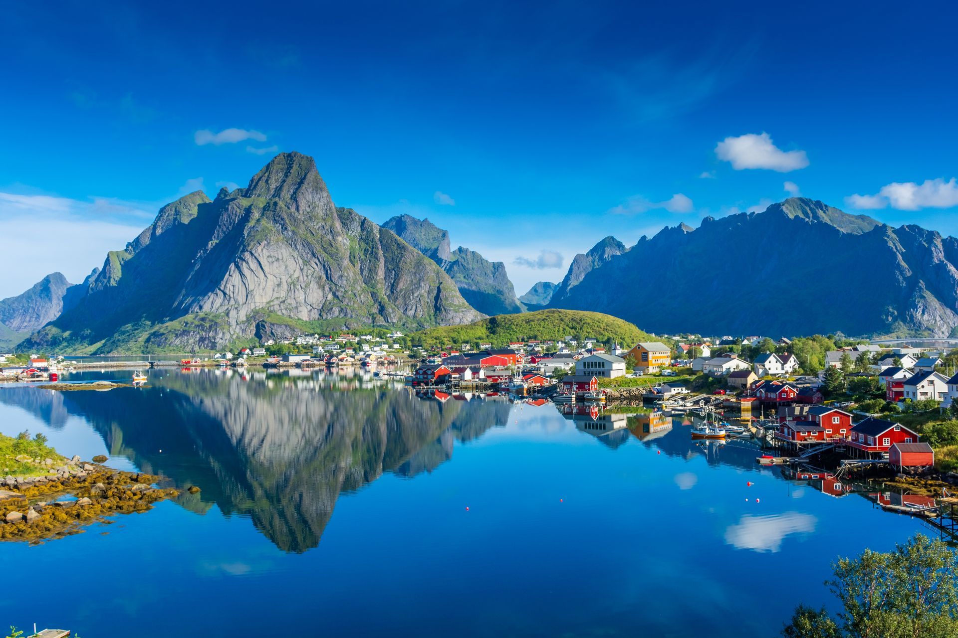 Mountains overlook a village with colorful buildings reflected in the calm water under a blue sky.