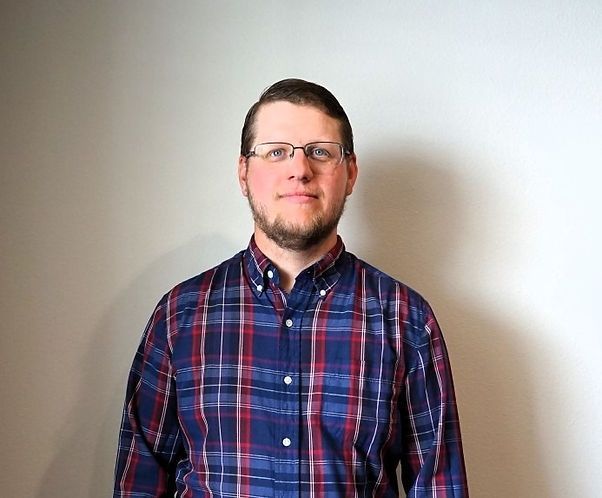 A man wearing glasses and a plaid shirt is standing in front of a white wall.