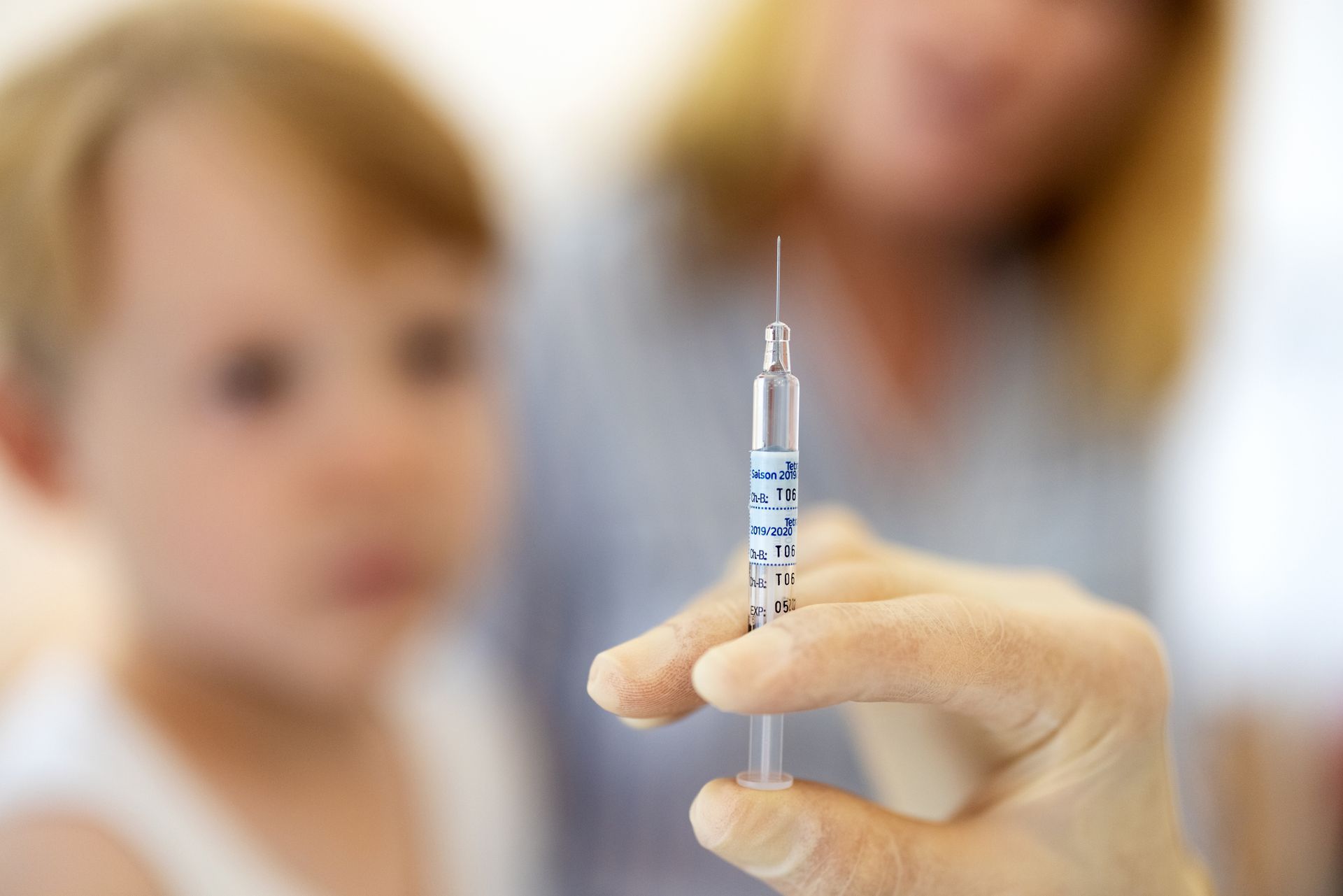 The doctor prepares a vaccine syringe as a young boy waits in the clinic background The doctor prepares a vaccine syringe as a young boy waits in the clinic background