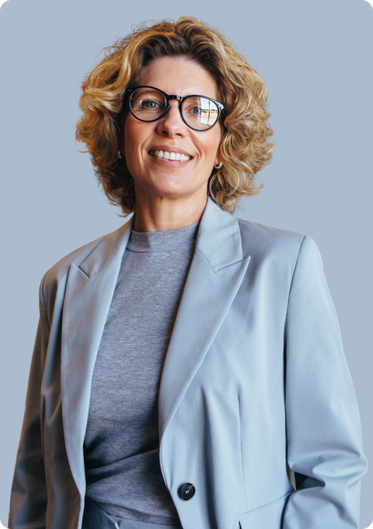 Woman wearing glasses, smiling, in a light blue blazer, grey shirt.