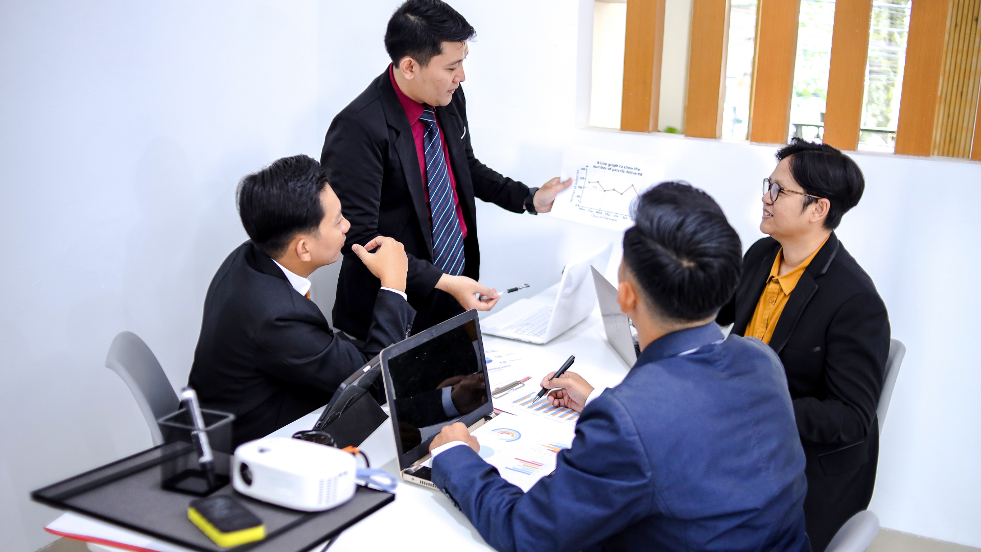 Business meeting: Five men in suits around a table, one standing and presenting papers, others focused on laptops.