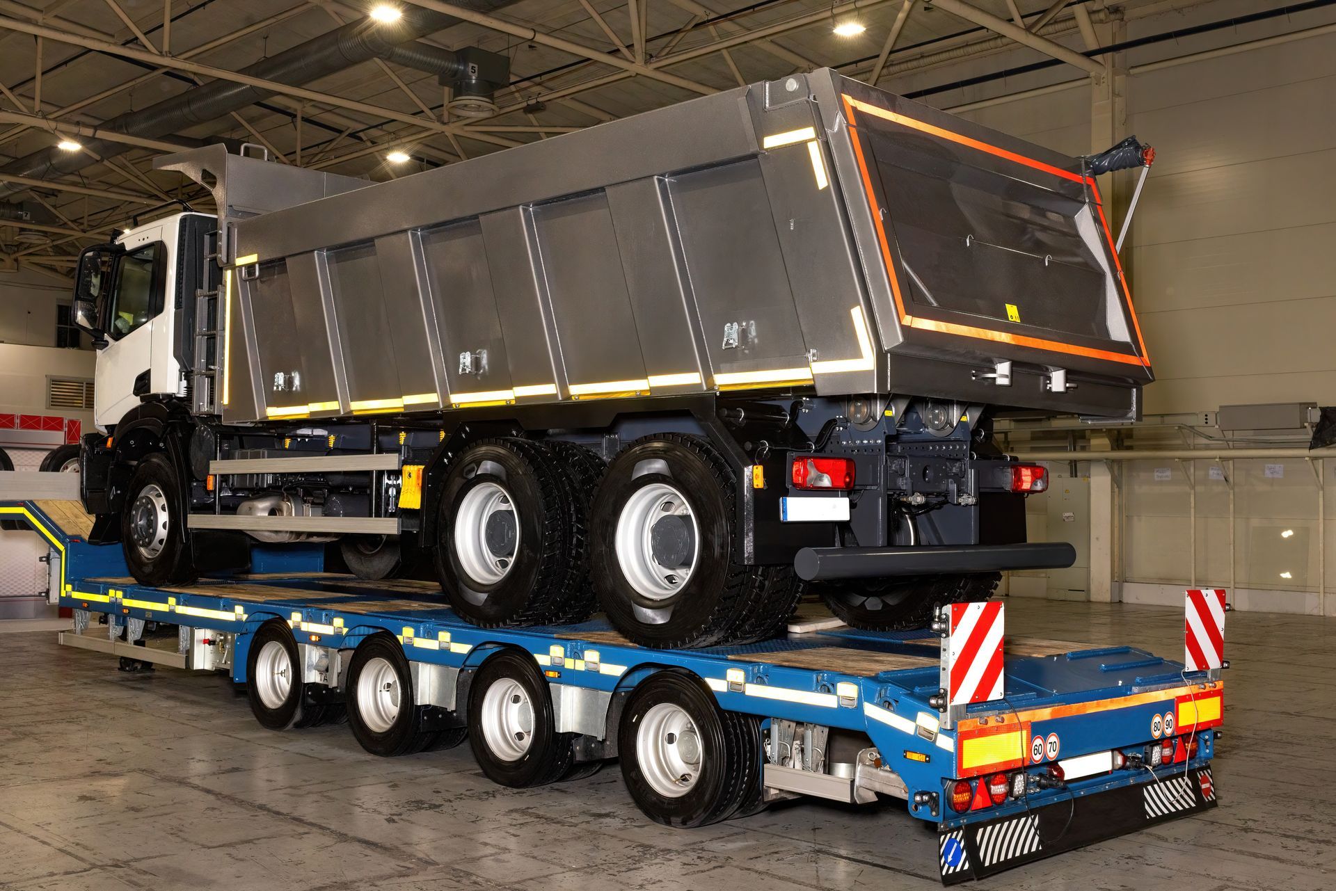 Dump truck on a blue trailer in a warehouse. The truck is gray and white.