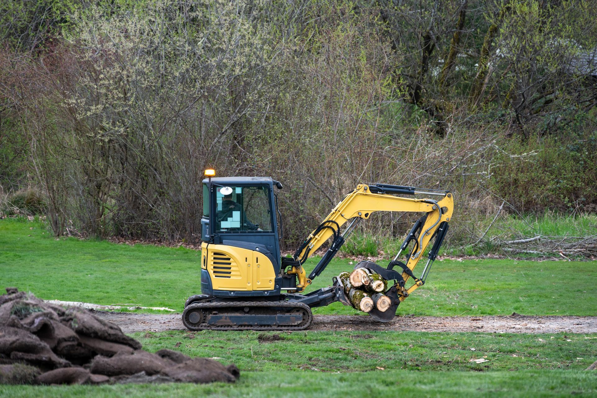 Yellow excavator in a grassy area, preparing soil. Trees in the background, a pile of earth on the left.