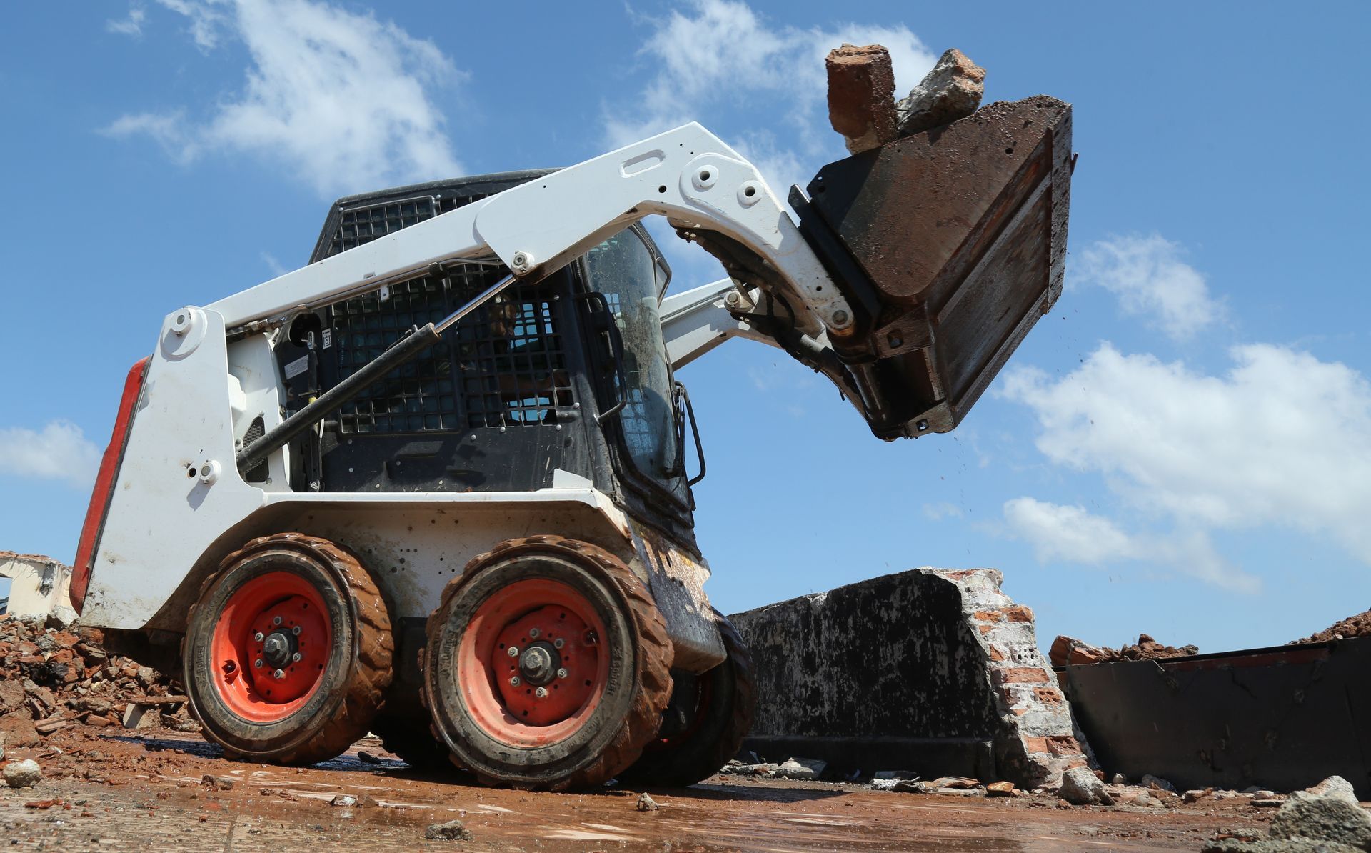 White Bobcat skid-steer loader scooping up debris at a construction site against a blue sky.