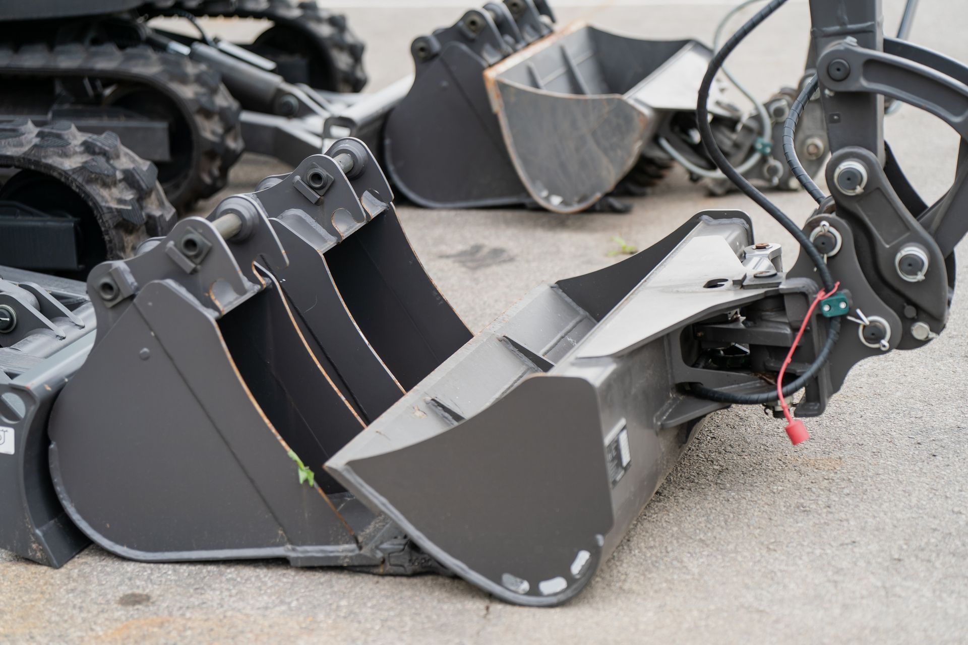 Excavator buckets in various sizes, gray metal, resting on pavement outdoors.