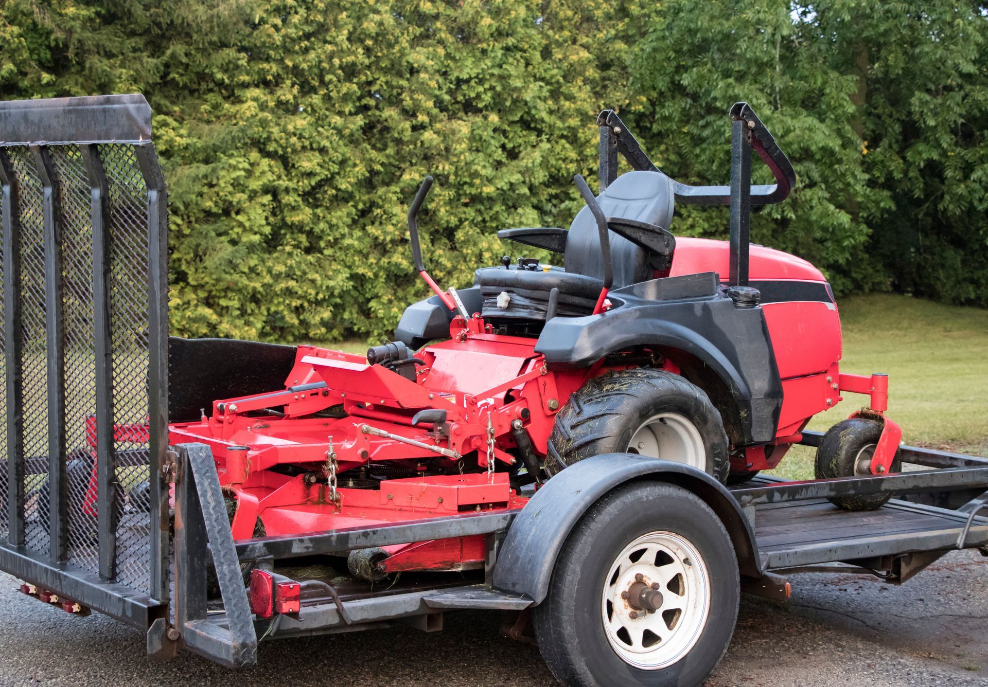 Red riding lawnmower on a trailer with a black cage, parked outdoors near green foliage.