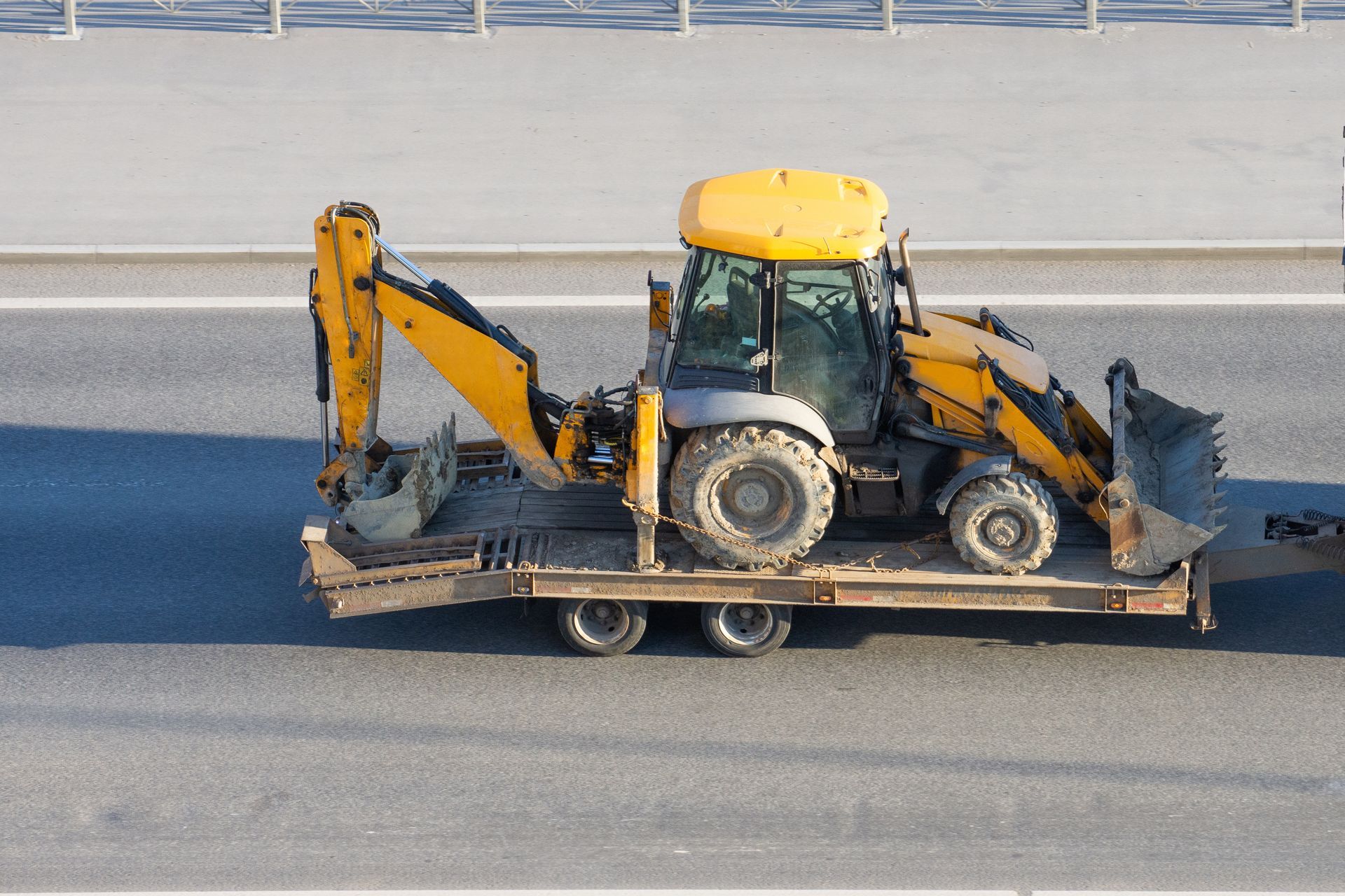 Yellow backhoe on a flatbed trailer, traveling on a road.
