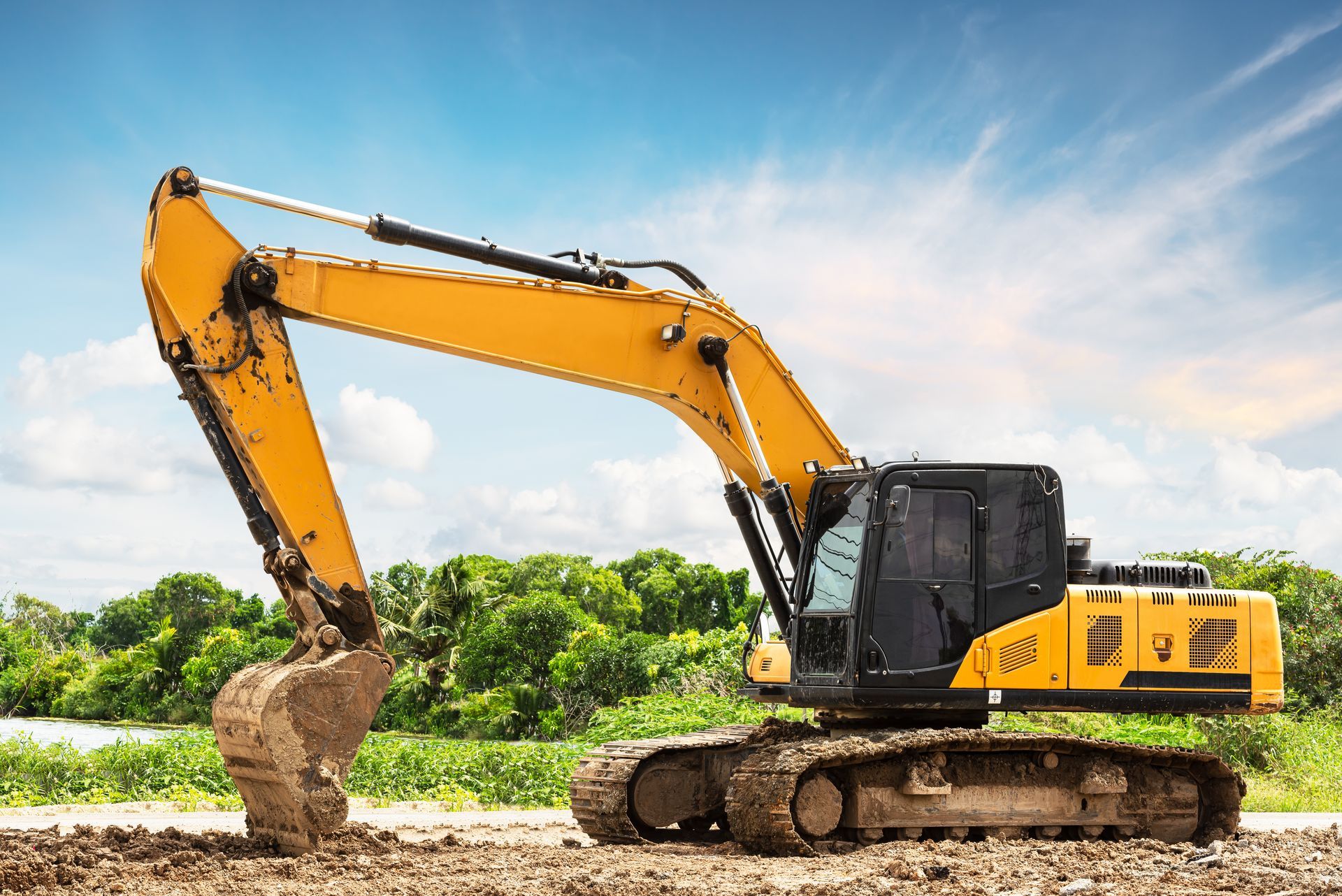 Yellow excavator digging on construction site, blue sky, trees in background.