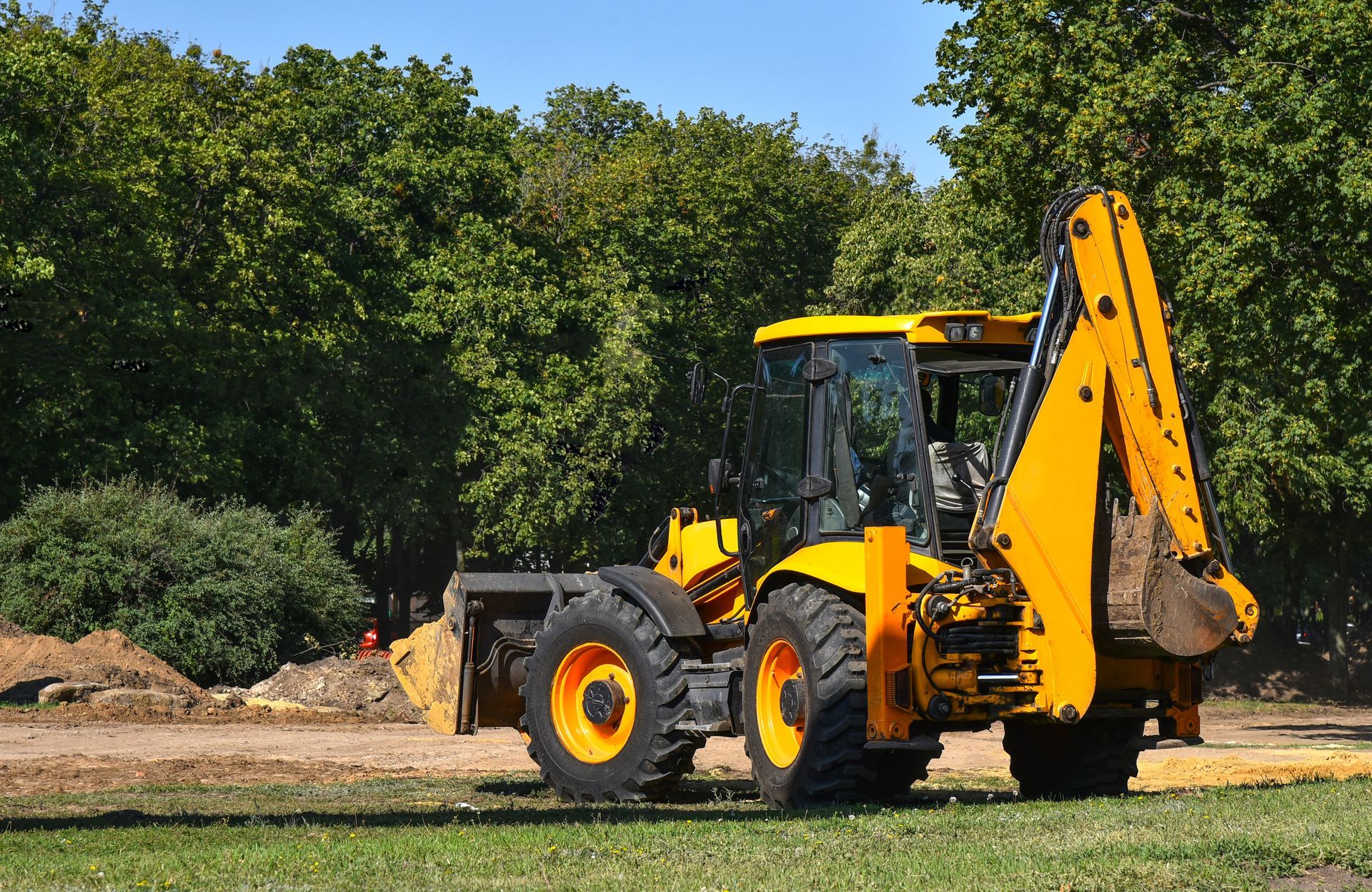 Yellow backhoe excavator on a grassy area, trees in the background, daytime.