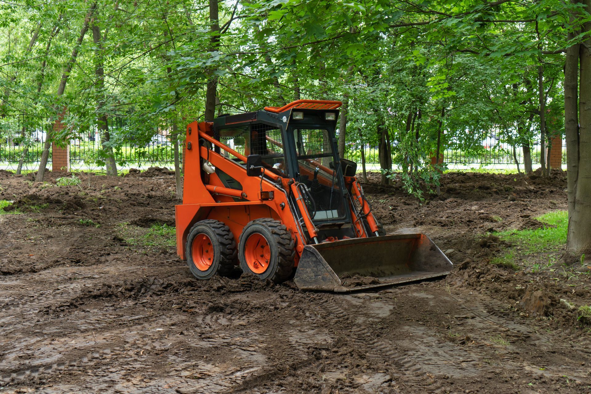 Orange skid steer loader on muddy ground in a wooded area.