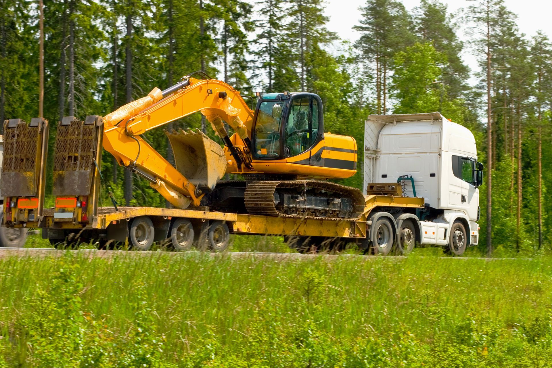 Yellow excavator on a flatbed truck traveling on a road, with trees in the background.