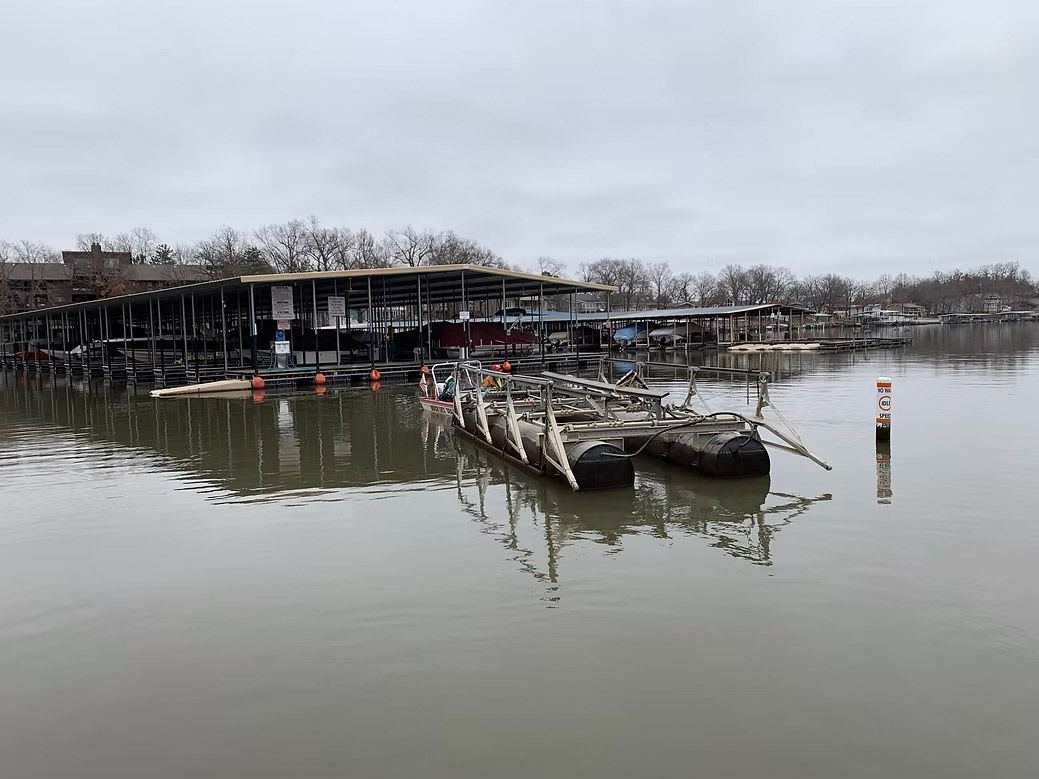 Flooded waterfront building with damaged docks reflecting in murky water under a cloudy sky.