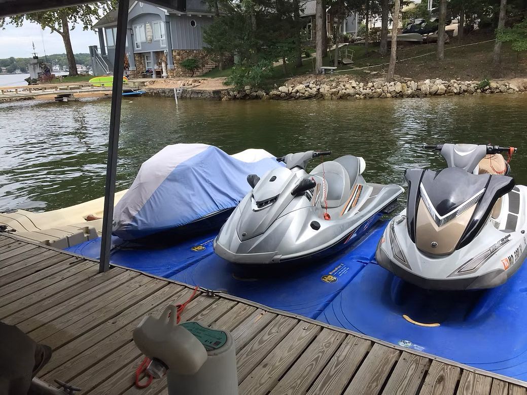 Three jet skis on a blue floating dock at a lake. One covered with a blue tarp, another silver, and one gray.