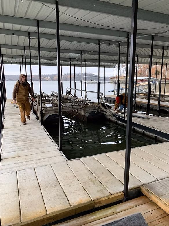 Man walks along a dock with another person working on a structure in a boathouse.