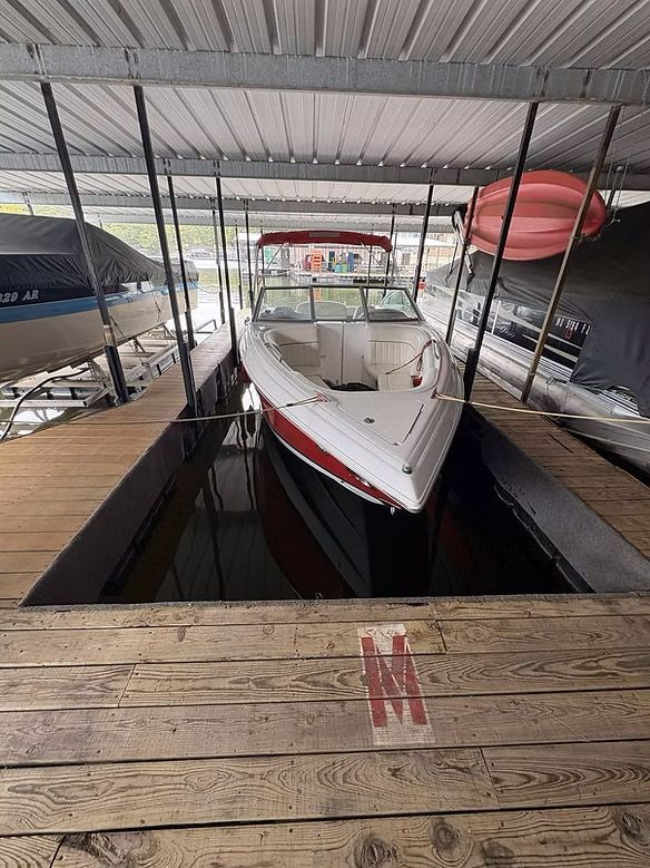 Boat docked in covered slip. White hull with red stripe. Dock with weathered wood, metal roof.
