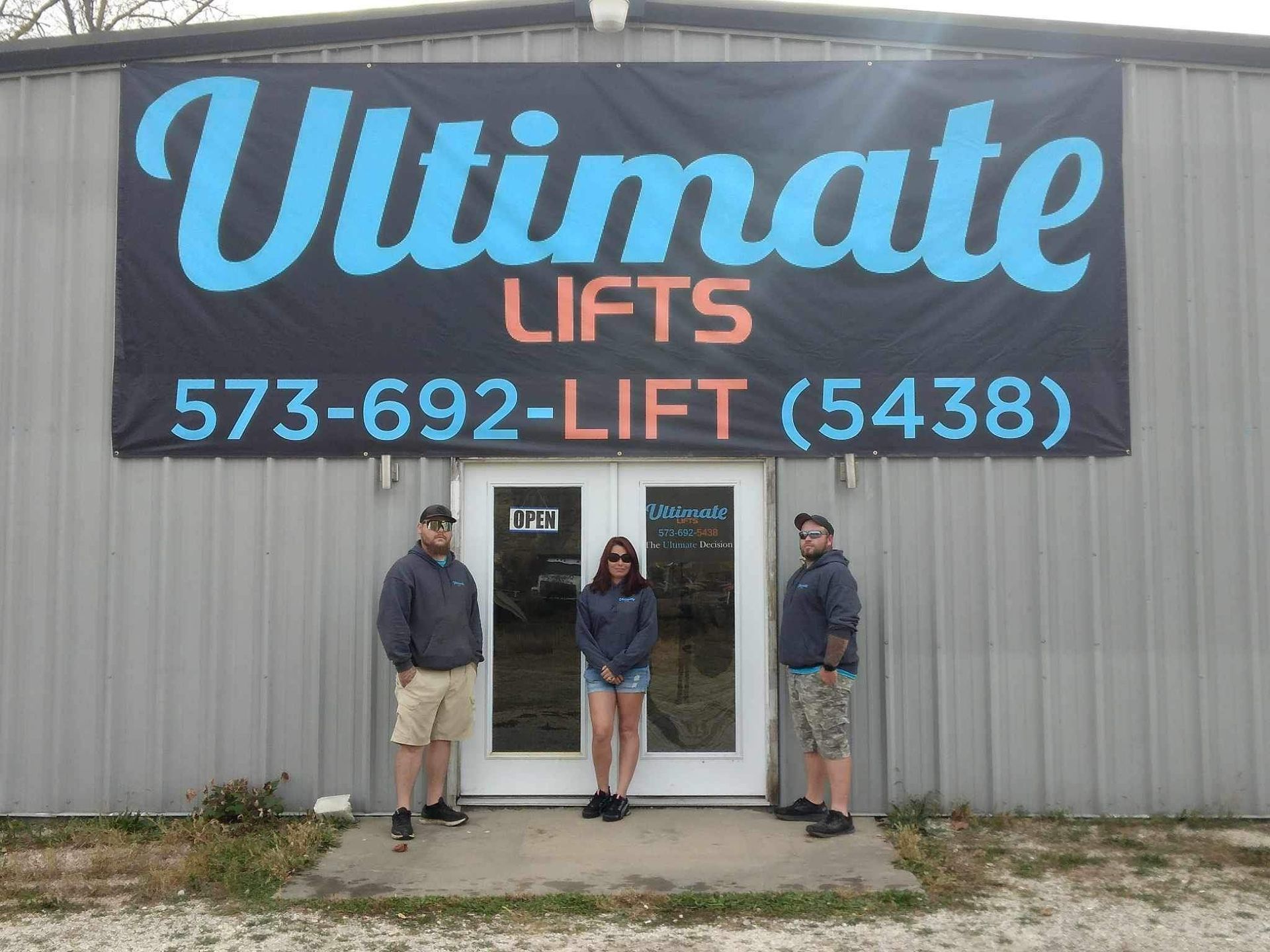 Three people stand in front of Ultimate Lifts building. Banner has business name and phone number.