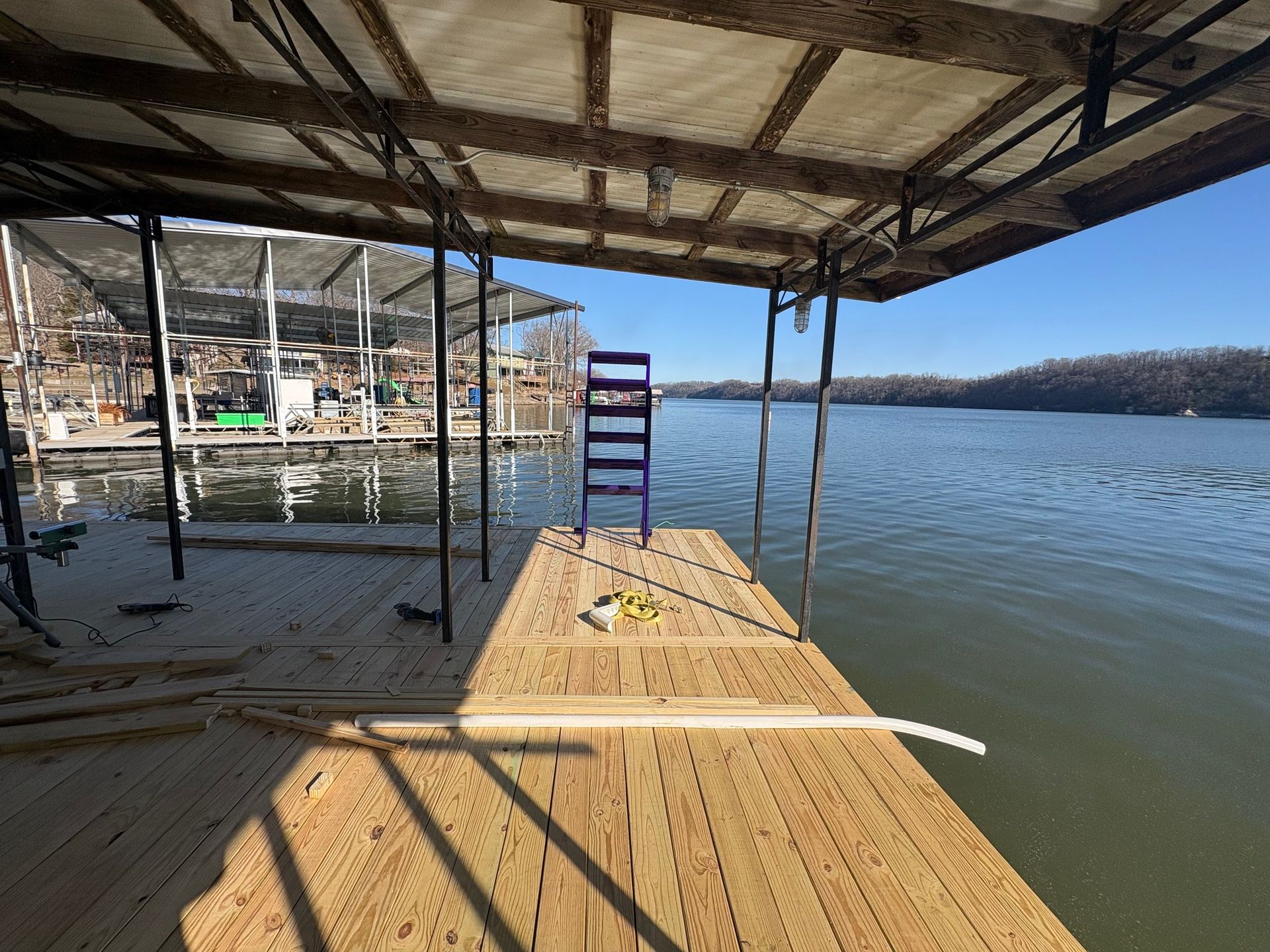 Dock overlooking a lake with a blue ladder, lumber, and protective roofing. Bright sunny day.