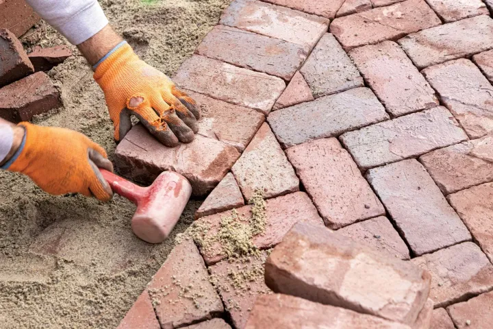 Gloved hands laying red brick pavers on sand with a rubber mallet nearby