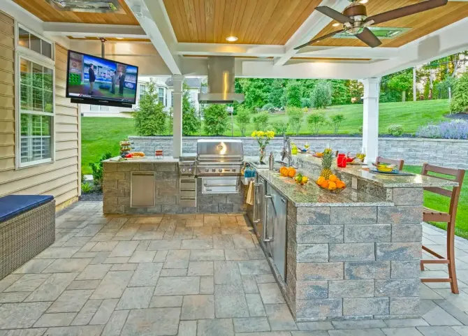 Covered outdoor kitchen and bar with stone counters, grill, TV, and patio seating under a wood سقف.