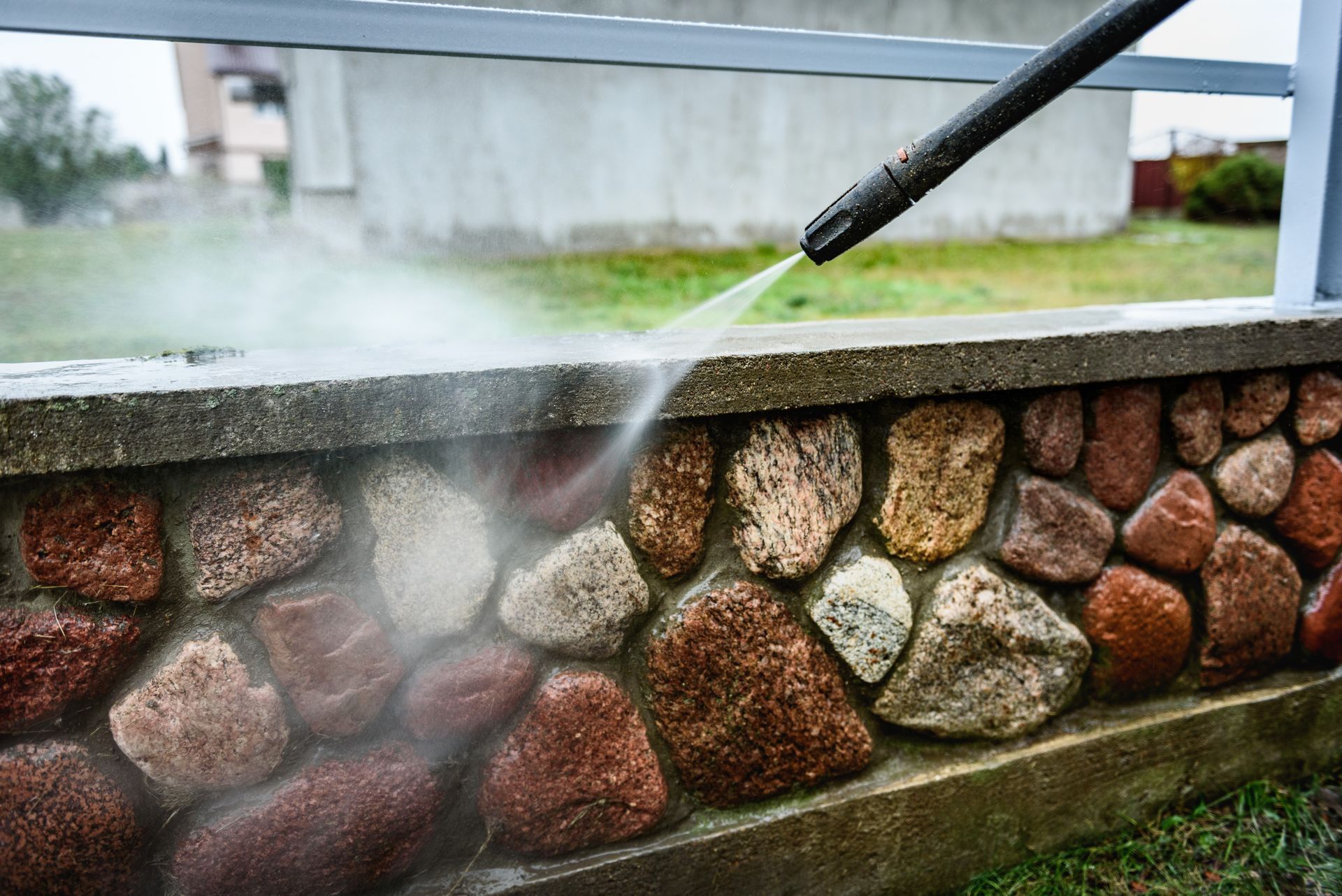 Close-up of a pressure washer spraying water to clean the top of a stone and concrete wall outdoors.